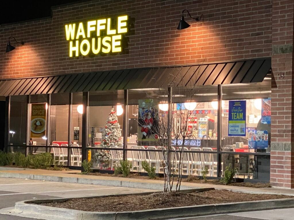 A well-lit Waffle House restaurant at night, featuring a glowing yellow sign above, Christmas decorations visible through the window, and a clear view of the interior from the outside.