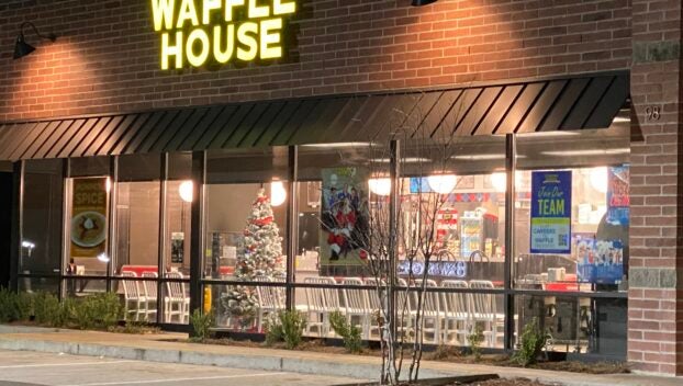 A well-lit Waffle House restaurant at night, featuring a glowing yellow sign above, Christmas decorations visible through the window, and a clear view of the interior from the outside.